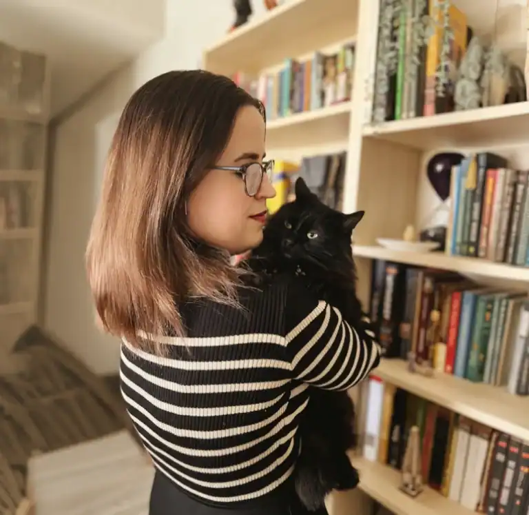 Daniella Marie, founder of Daniella Marie Beta Reading Services, holding her cat Toothless in front of her personal bookshelf at home in Florida.